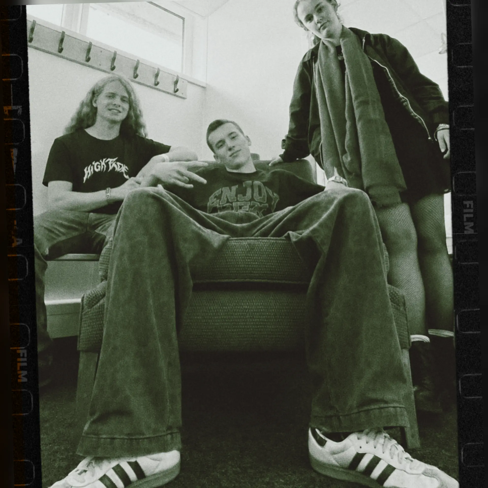 Black and white film-strip portrait of Funky Biscuit in a dressing room, all three members clearly visible posed around an armchair with coat hooks on the wall behind them