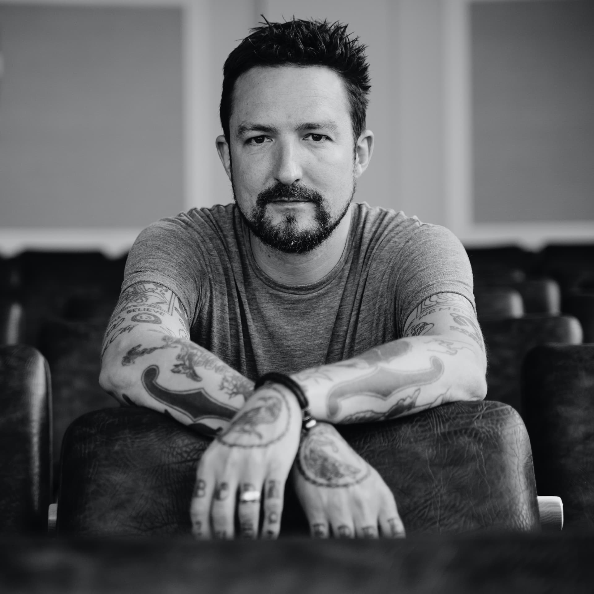 Black and white press portrait of Frank Turner seated with his tattooed arms crossed